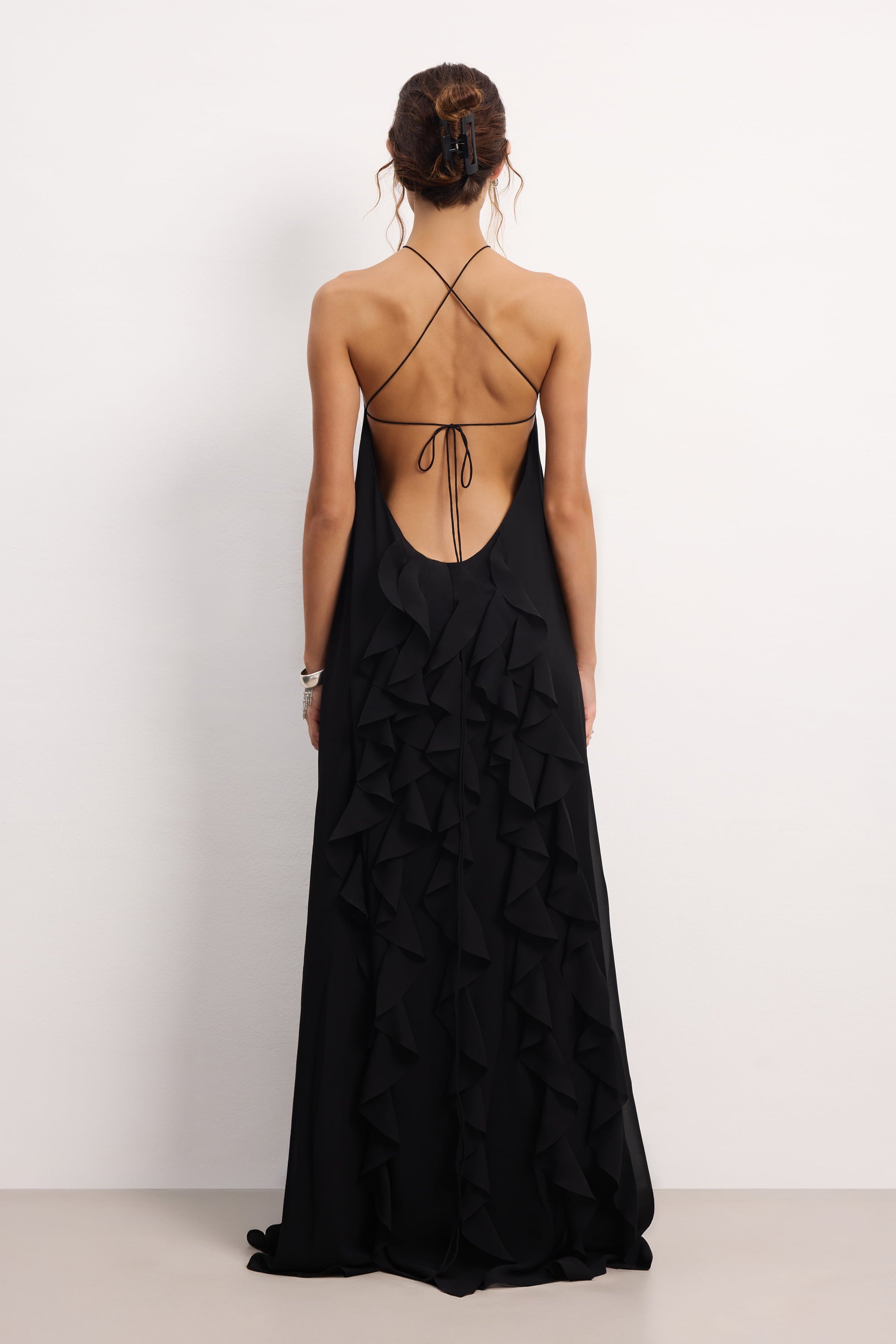 Woman wearing a black halter neck Illusion dress with thin straps and ruffled layers on a white background, back view