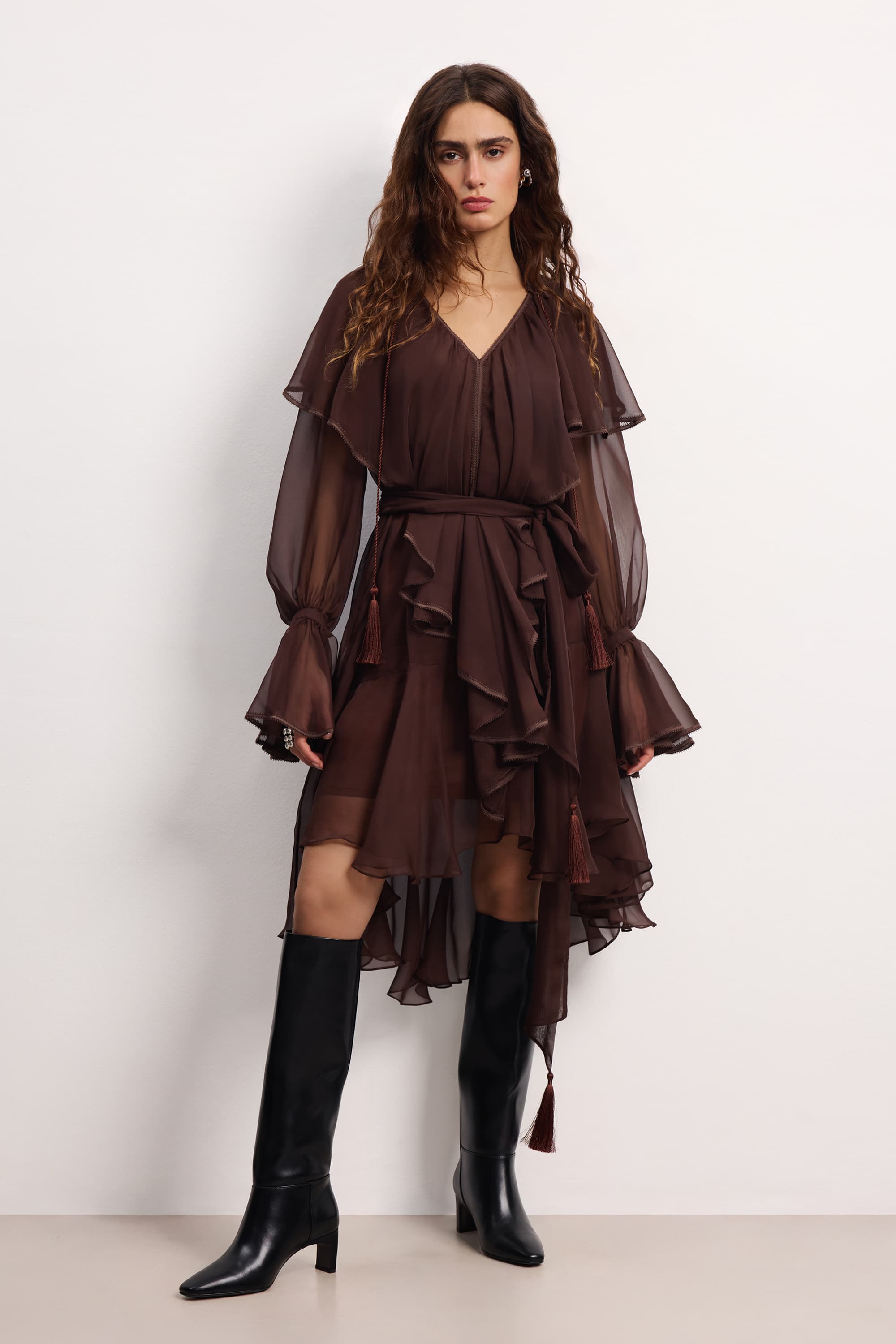 Woman wearing a brown ruffled Ocean Of Tenderness midi dress with black knee-high boots against a white background, front view