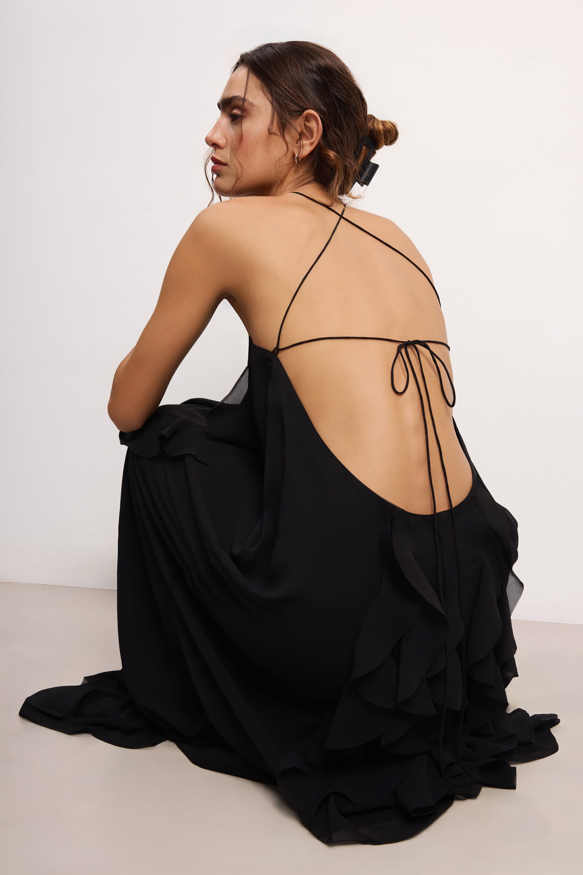 Woman wearing a black halter neck Illusion dress with thin straps and ruffled layers on a white background, back view