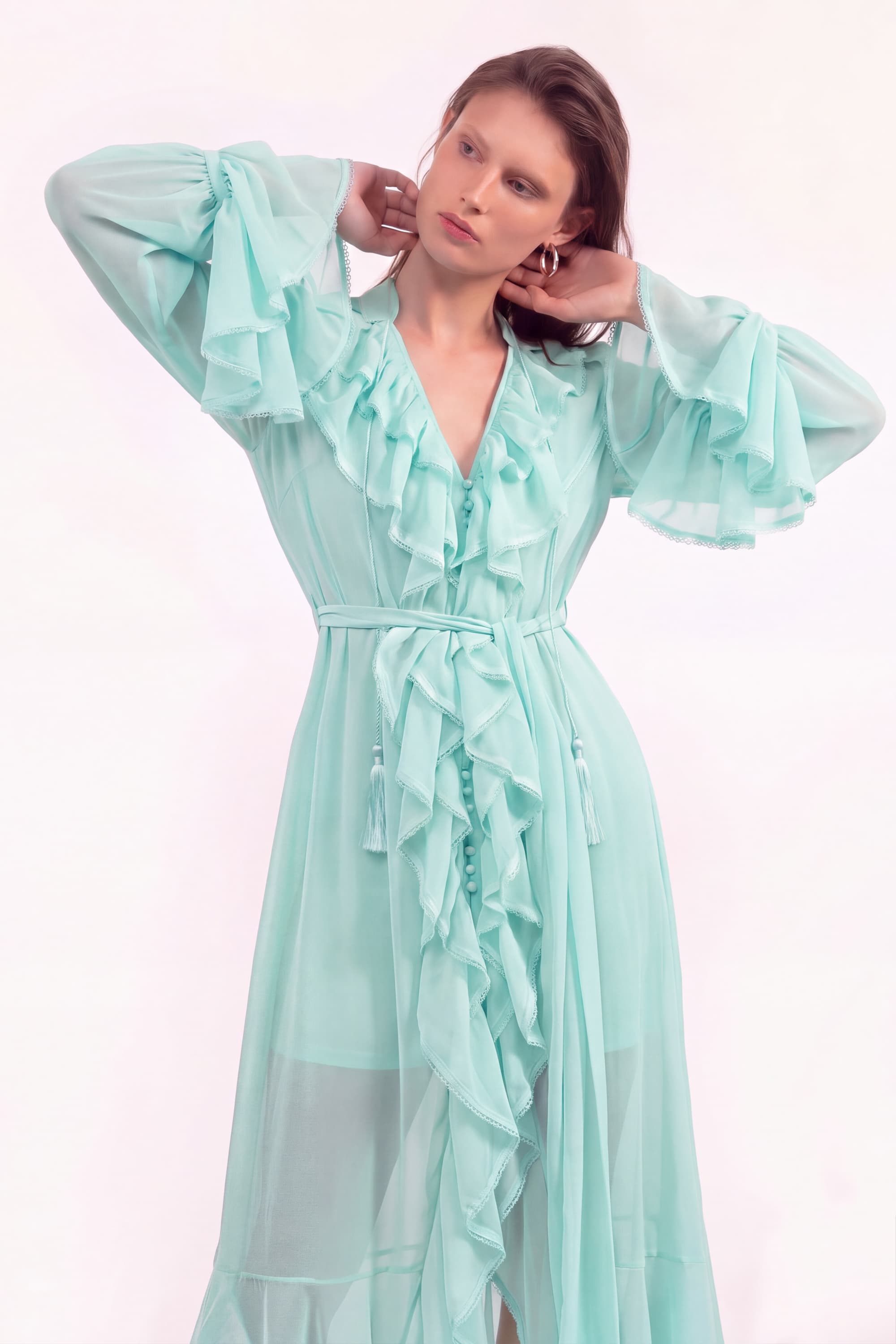 A woman modeling a long, flowing mint green dress with dramatic ruffled sleeves, a deep V-neckline, and a matching ruffled tie belt at the waist, set against a plain light background.