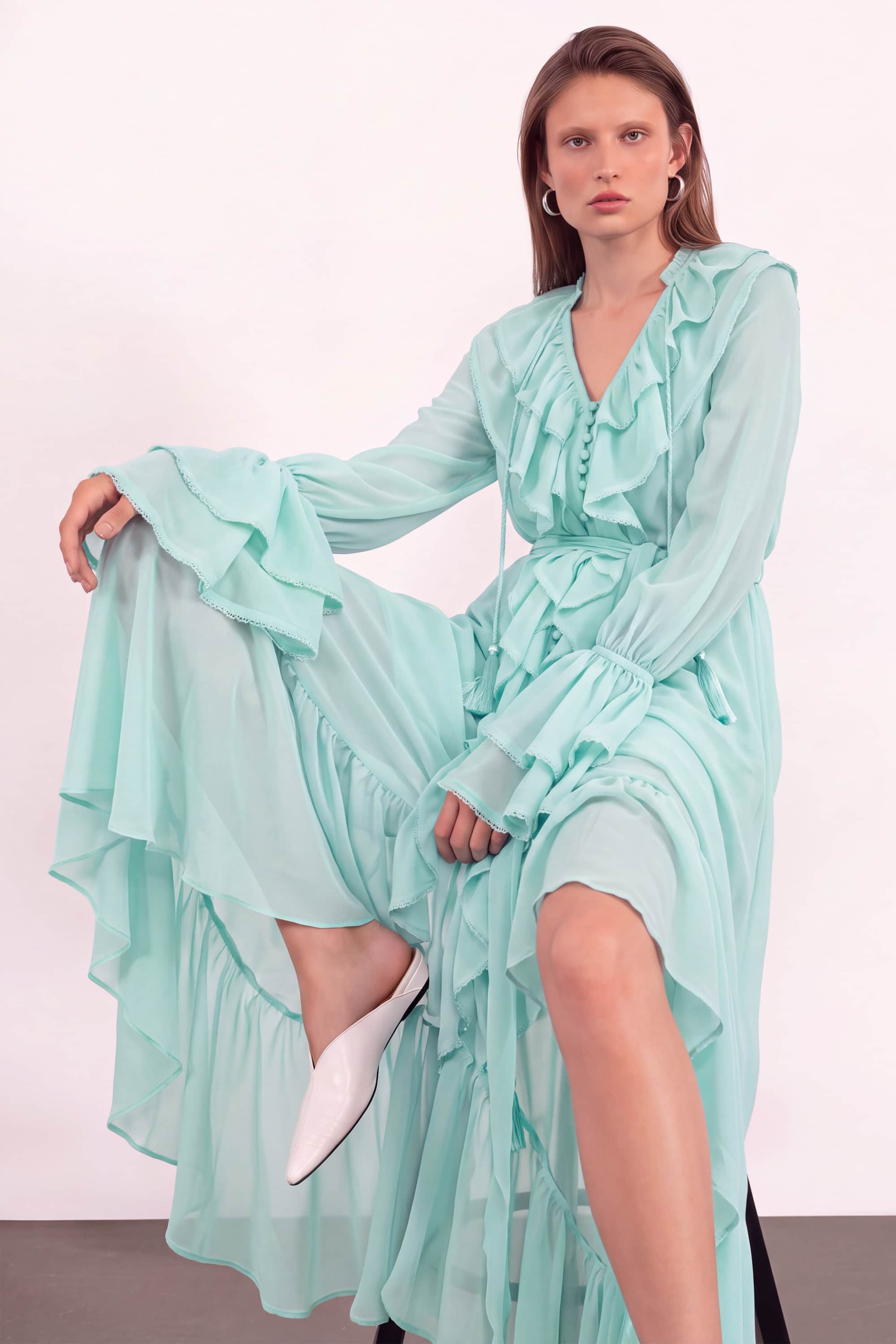 A woman models a long-sleeved, ruffled mint green dress with a high-low hemline and white pointed-toe flats, posed against a plain off-white studio background.