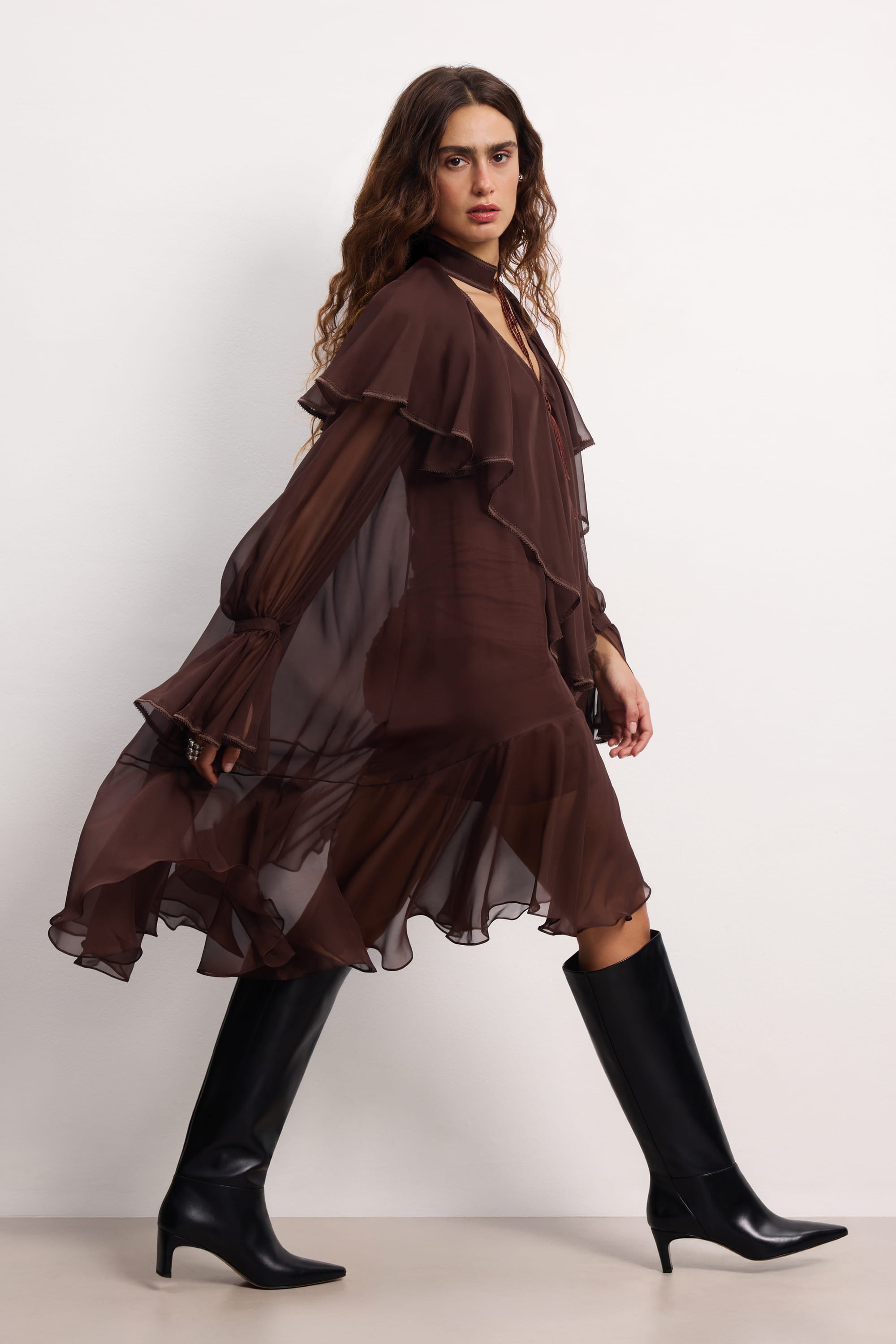 Woman wearing a brown ruffled Ocean Of Tenderness midi dress with black knee-high boots against a white background, side view