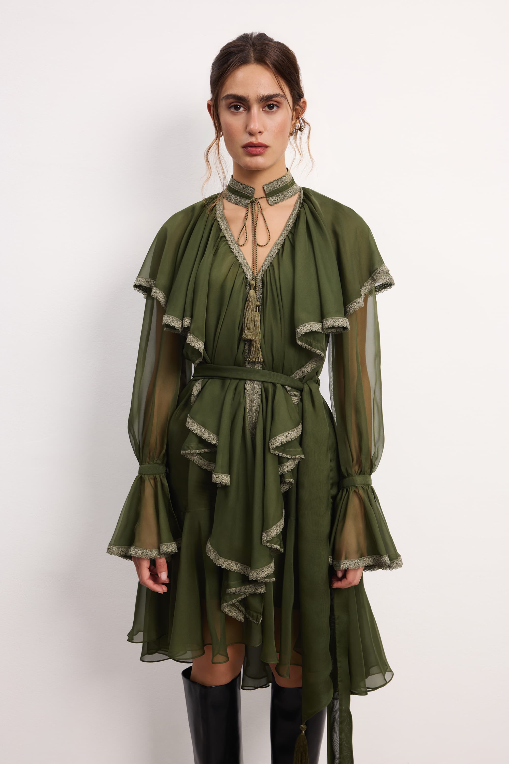 Woman wearing a olive ruffled Ocean Of Tenderness midi dress with black knee-high boots against a white background, front view